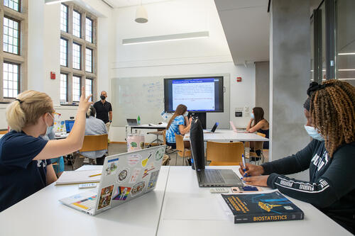 A student raises her hand during a class in a Poorvu Center classroom. Other students are working at their laptops, and the professor looks to the student.
