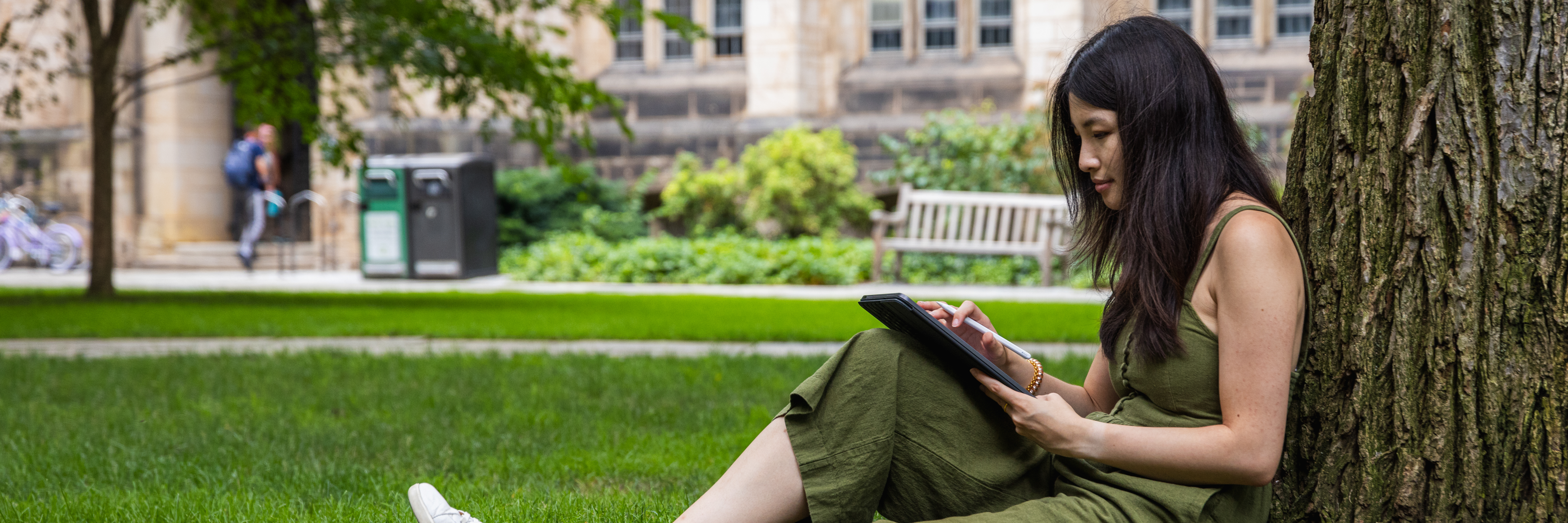 A student on Cross Campus writing on a tablet sitting on the grass and leaning up against a tree.