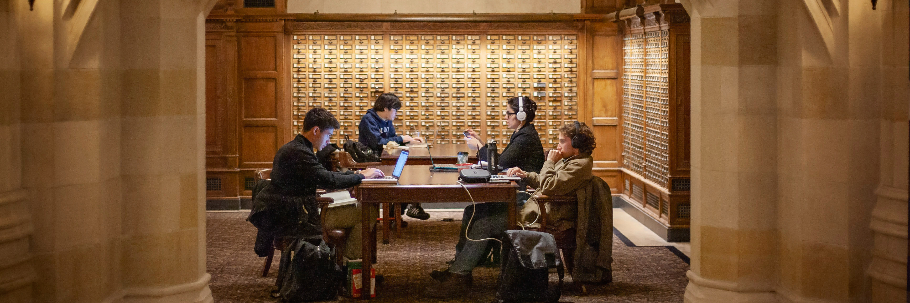 Four grad students writing on laptops at a table near the old card catalog drawers in SML