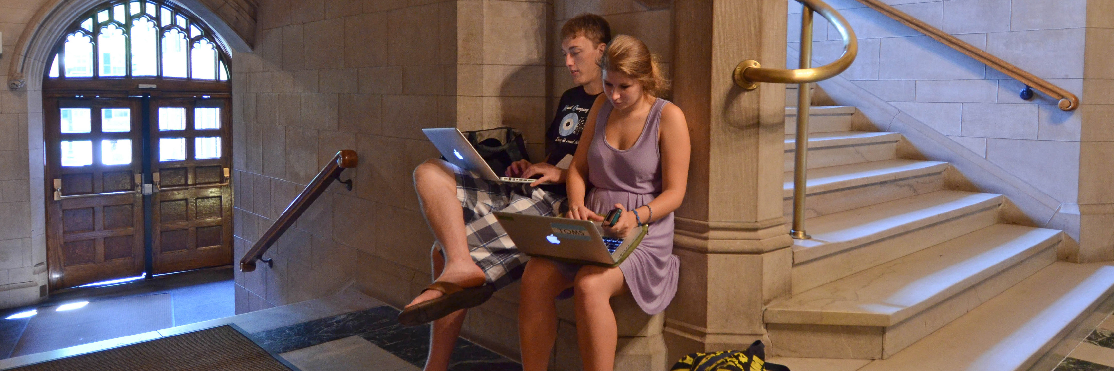 Two students writing on laptops near a stairwell in Linsley-Chittendon Hall