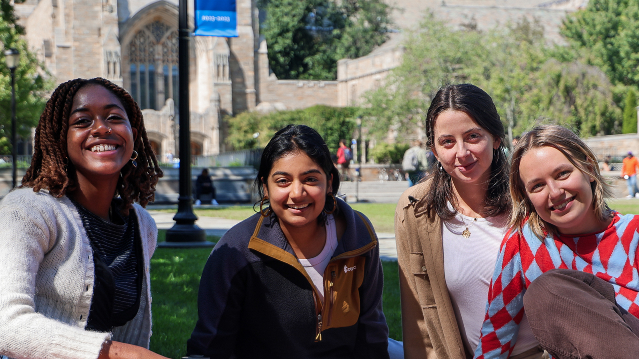 Four students sitting on a blanket in front of Sterling Memorial Library