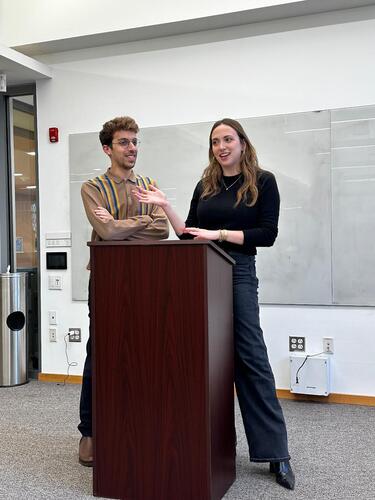 Ben and Rebecca stand behind a podium in a classroom, smiling and talking 