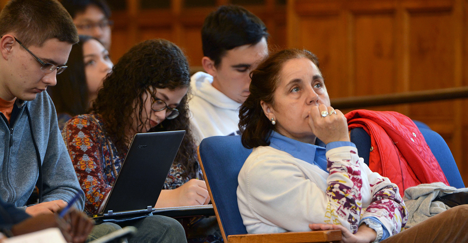 A Yale professor sits in a traditional classroom, listening attentively. She is surrounded by students taking notes.