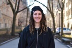 A person with long curly hair wearing a black jacket and hatDescription automatically generated