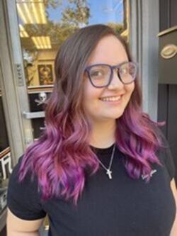 A person with purple hair and glasses smilingDescription automatically generated