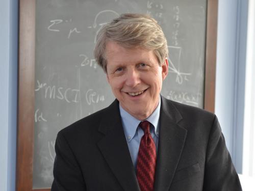 Professor Robert Shiller, wearing a suit with a red tie and blue shirt, stands in front of a black board.