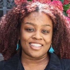 Micah Nellums in a black collard shirt standing against a floral background.