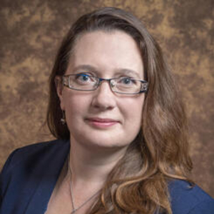 Pilar Abuin standing in a blue blouse and sweater on a brown background.