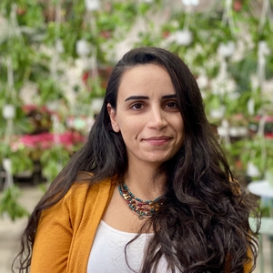 Yaprak sits outside and smiles slightly at the camera. She wears a white shirt, an orange cardigan, and a necklace. She has long dark hair.