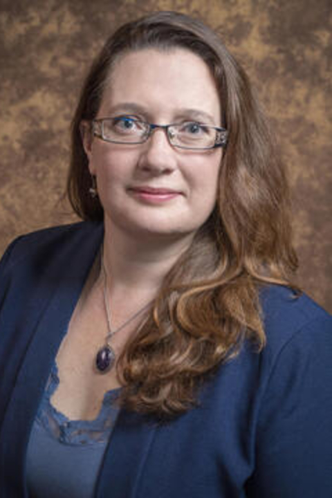 Pilar Abuin standing in a blue blouse and sweater on a brown background.