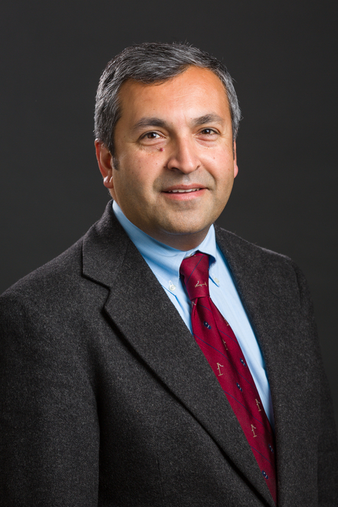 Professional photo of Mayur Desai, a man wearing a dark suit jacket, light blue shirt, and red tie with small patterns. He has short, salt-and-pepper hair and is smiling slightly, posed against a plain dark background.