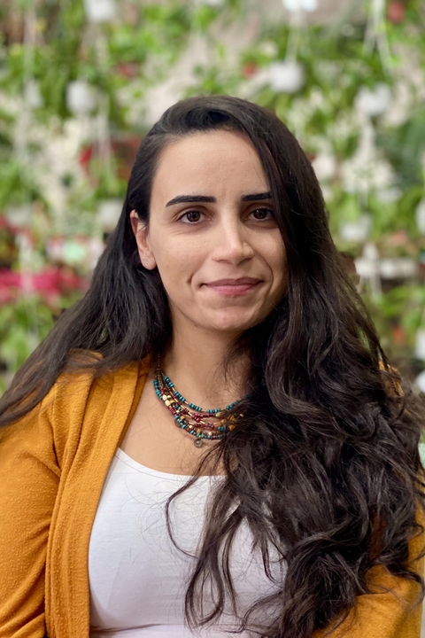 Yaprak sits outside and smiles slightly at the camera. She wears a white shirt, an orange cardigan, and a necklace. She has long dark hair.
