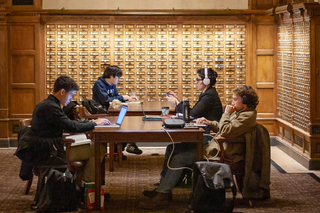 Four grad students writing on laptops at a table near the old card catalog drawers in SML