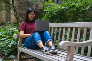 A student smiles at their laptop while sitting on a bench outside.