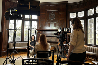 Behind a camera for a recorded interview. Two people sitting for the interview with camera lights set up to the side in a rich dark wood interior room.
