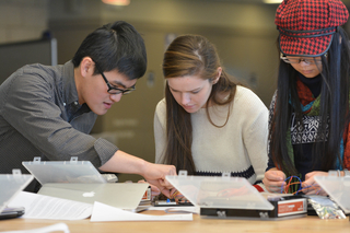 Three students sit at a table working with an electronics project.