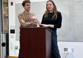 Ben and Rebecca stand behind a podium in a classroom, smiling and talking 