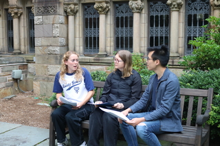 Three students exchanging feedback on a paper on a bench in a Yale courtyard.