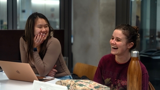 Two Writing Partners sit at a table with a laptop open. They are smiling and laughing.