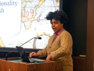 A young woman stands at a podium speaking. A map of the world is visible on a screen behind her.