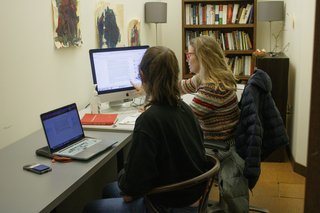 A woman sits at a desk and points at her computer while a student at her side looks on.
