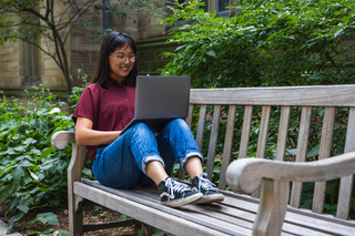 A student smiles at their laptop while sitting on a bench outside.