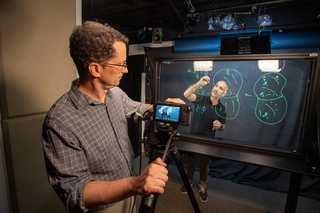 John Harford filming teacher writing on transparent board.