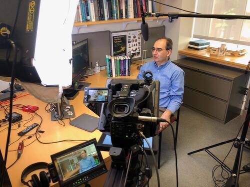 Professor Xenophon Papademetris sits at his office desk surrounded by professional lighting and speaks to a video camera